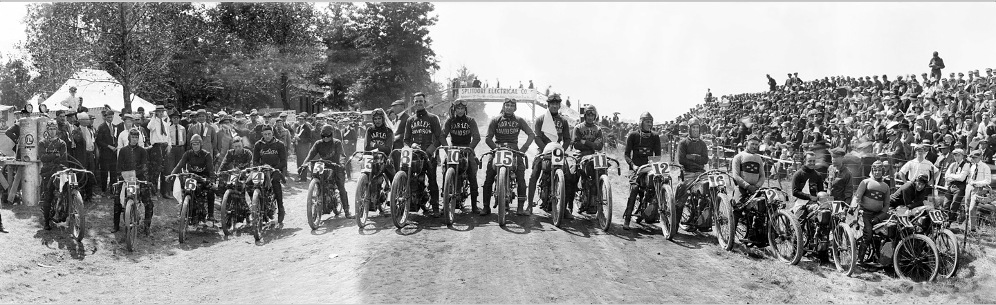 Original black-and-white 1919 panorama: eighteen motorcycles and riders across the dirt road, pits and trees on the left, packed bleachers on the right, and the Splitdorf bridge behind.
